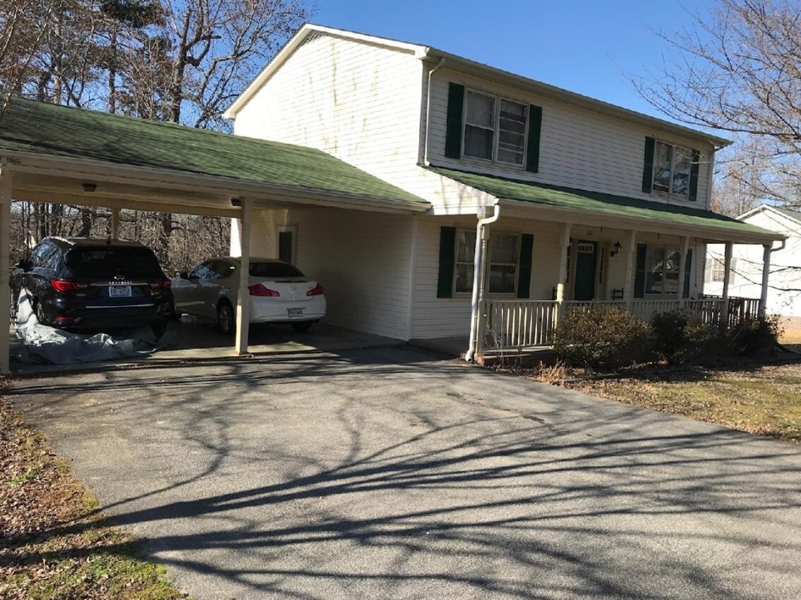 Two‑story house with green shutters and a carport, two cars parked in the driveway, and bare winter trees casting shadows across the yard belonging to Sponsored Residential Provider Armando Cooper in Danville, Virginia.