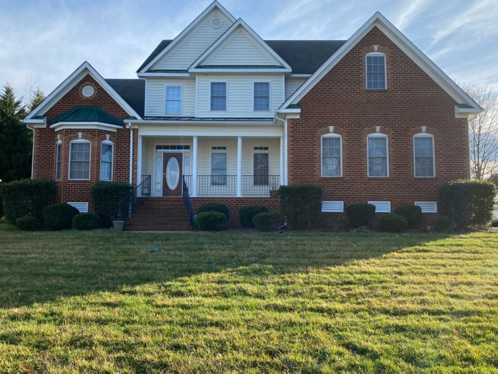 Two‑story brick and white‑siding house with gabled rooflines, a covered front porch with columns, and neatly trimmed shrubs in front, set on a grassy lawn belonging to Group Home Providers Dayshawnna and Michael Lee in Chester, Virginia.