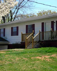 Single‑story white house with red shutters and a small wooden front porch on a grassy lawn belonging to Group Home Provider Donald Nester in Danville, Virginia.