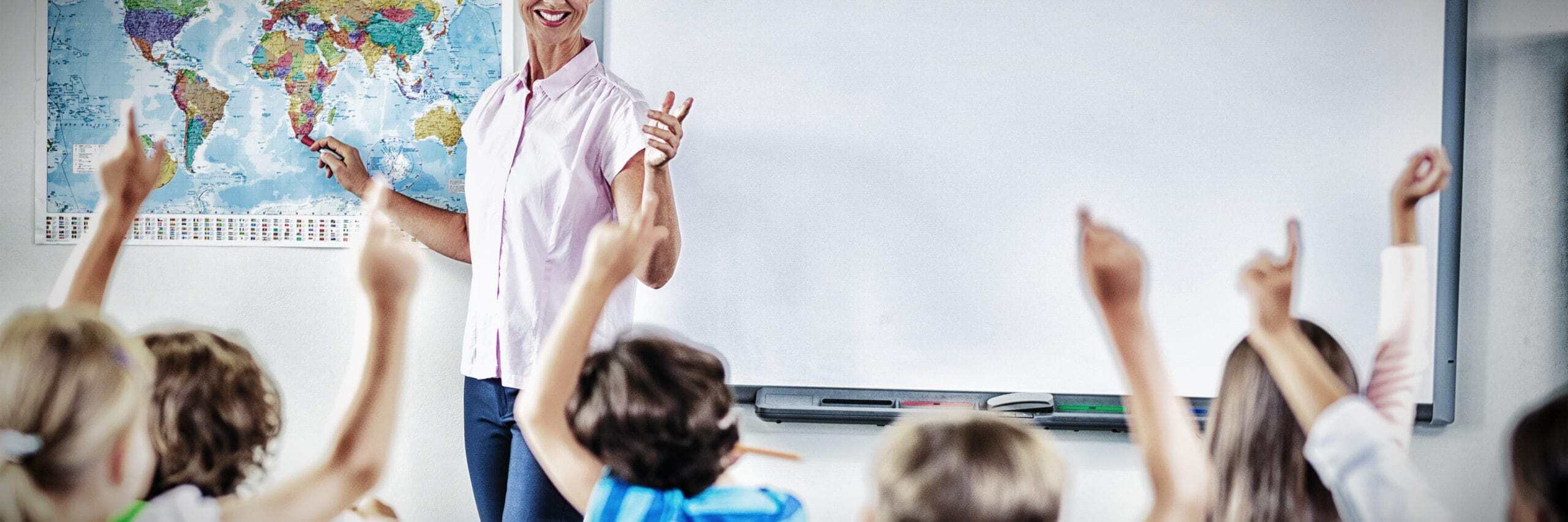 Classroom view from the back with children raising their hands while the teacher stands at the front