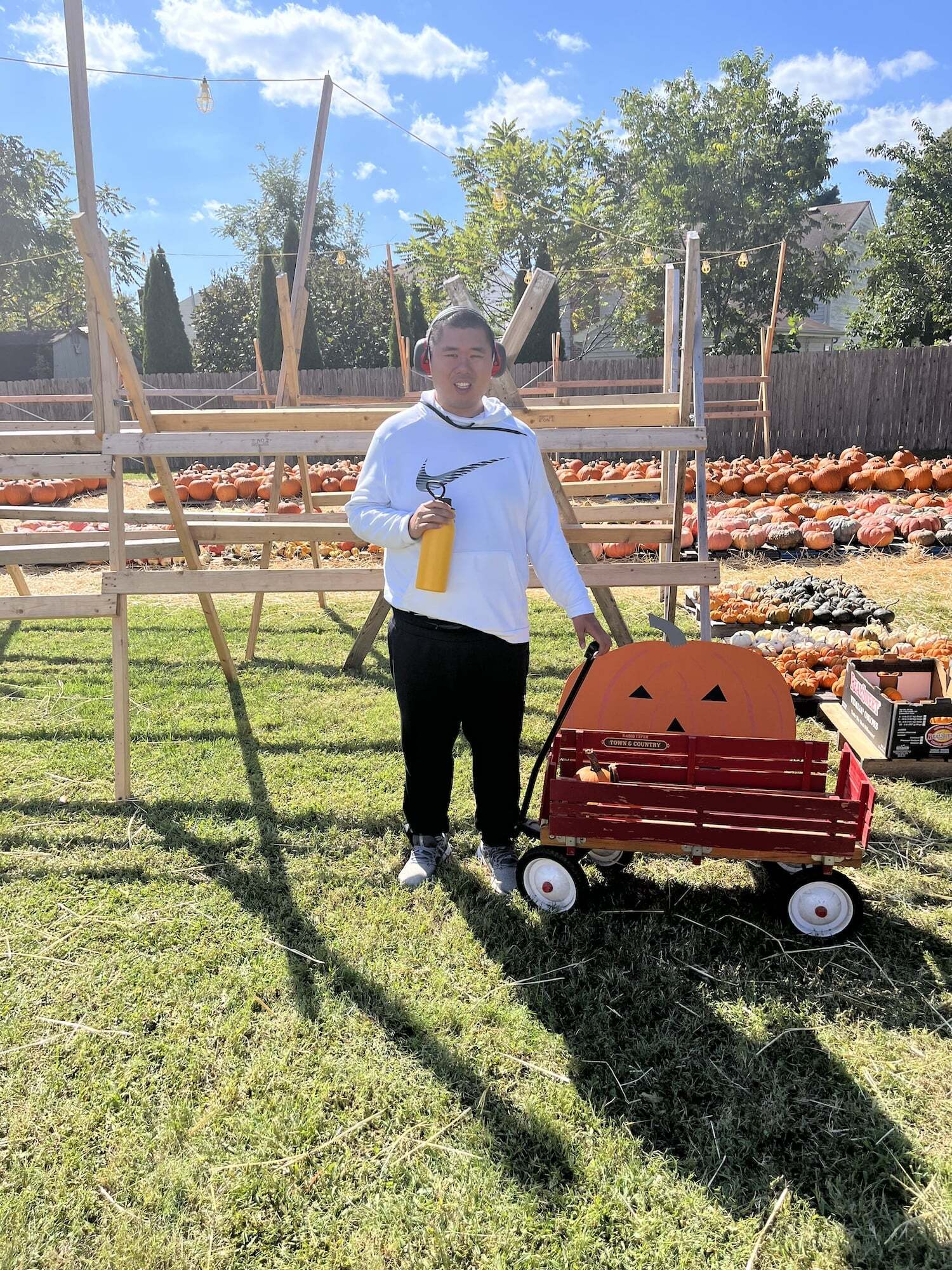 An individual in a white hoodie standing outside with a red wagon in a pumpkin patch on a sunny day