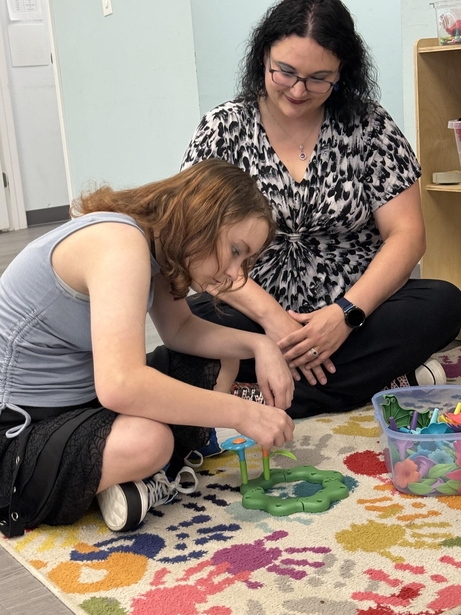 A woman and a girl sitting cross-legged on the floor playing with toys