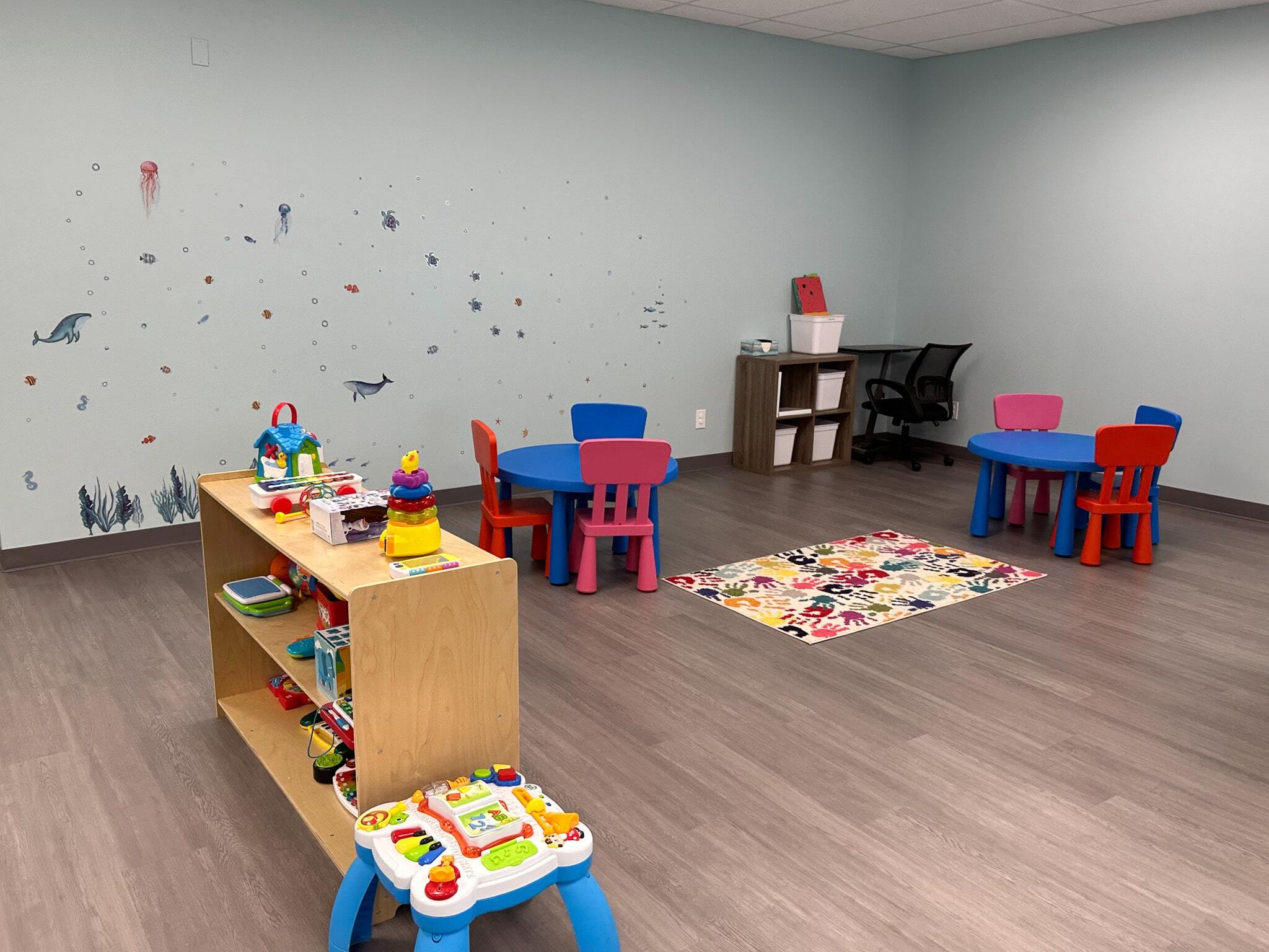 An open room with tables and chairs for children,  a desk in the corner, and toys and activities